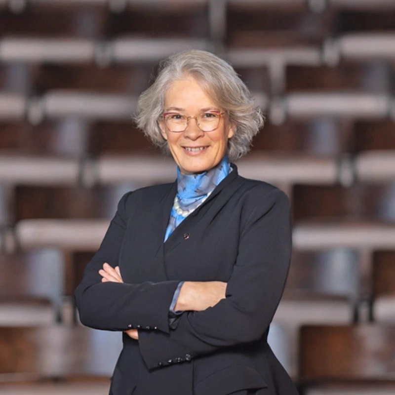 Smiling senior college woman with glasses and black blazer standing out in classroom or conference room.