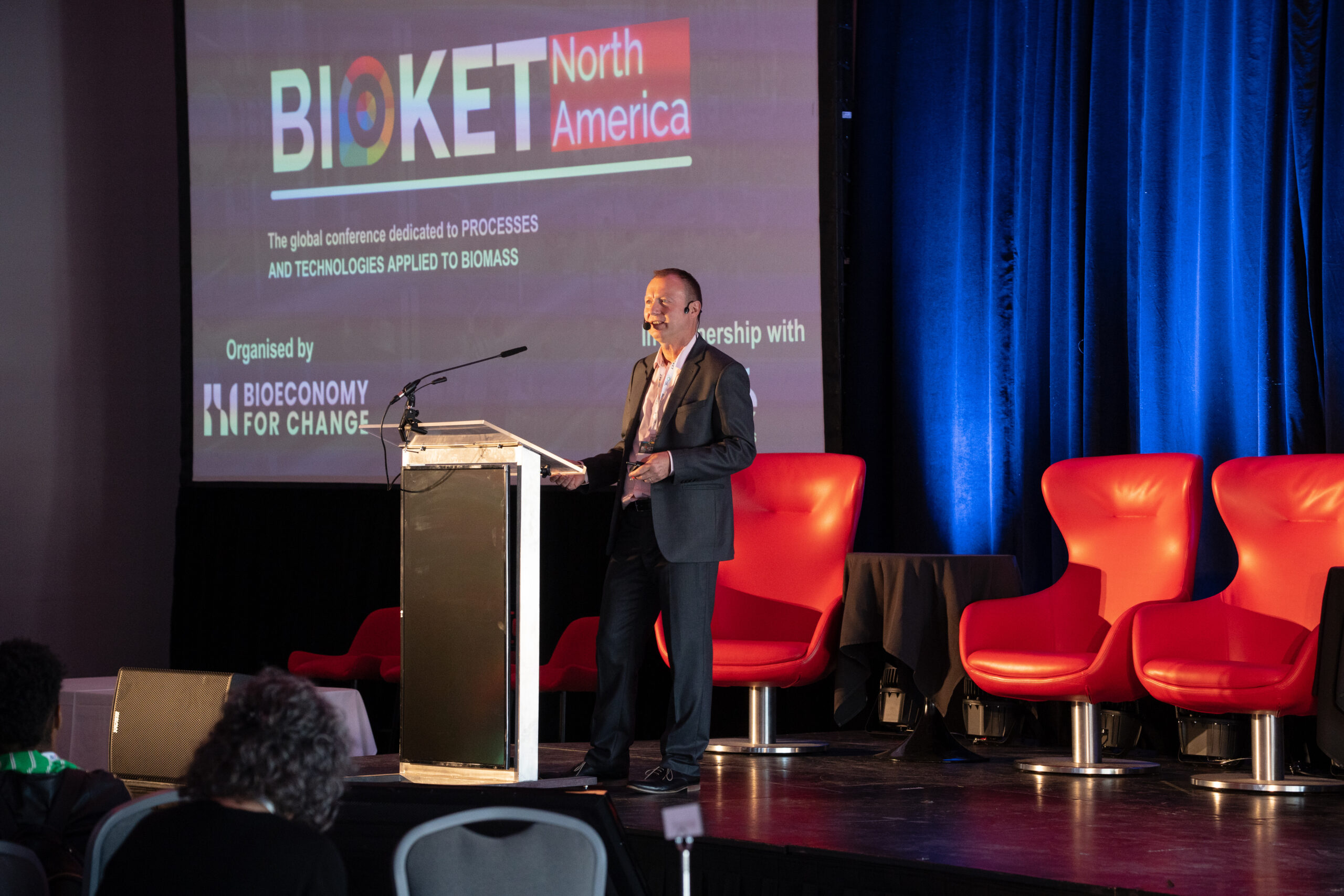 Palestrante apresentando no evento BioKET North America, que discute processos e tecnologias aplicadas à biomassa na conferência de bioeconomia, com palco equipado e assentos vermelhos.