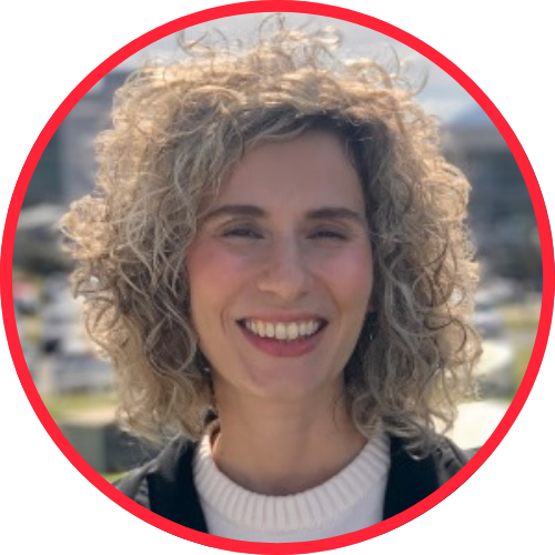 Retrato sorridente de uma mulher com cabelos cacheados loiros, em ambiente externo, com o sol iluminando o rosto e um círculo vermelho em destaque na imagem.