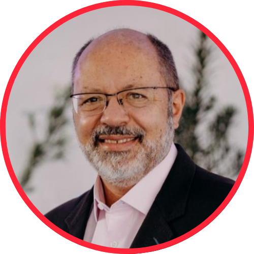 Retrato de homem de meia-idade com barba grisalha, óculos e terno escuro, sorrindo ao ar livre, com fundo desfocado e moldura circular vermelha